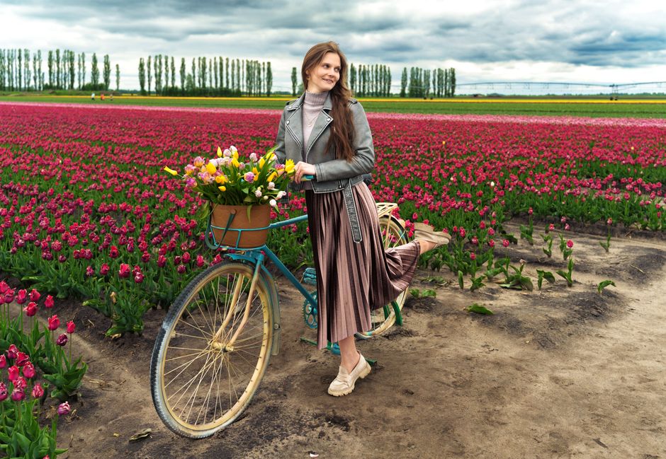 Woman stands next to a bicycle with flowers in a vibrant tulip field under cloudy skies.