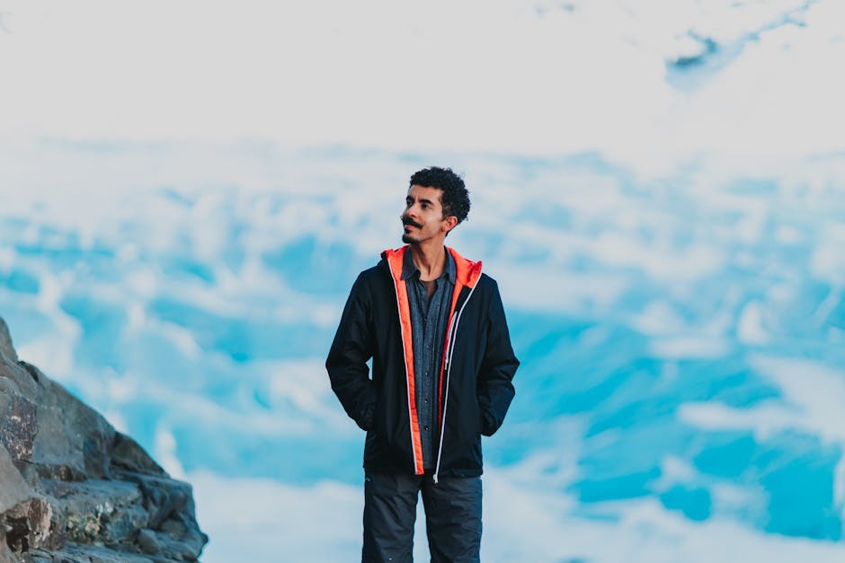 Content ethnic guy in warm winter jacket holding hands in pockets looking dreamily away while standing on cliff in nature