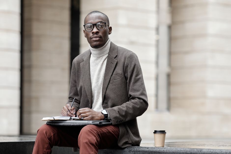 A professional man in a suit jacket sits outdoors, taking notes with a coffee cup beside him.