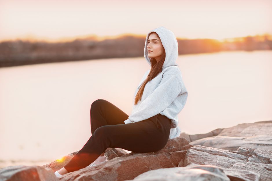 A serene scene of a woman in a hoodie sitting on rocks by the water during sunset.