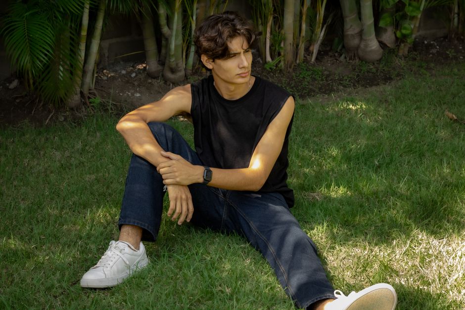 A young man in jeans and a sleeveless shirt sits thoughtfully on grass in a sunny garden.