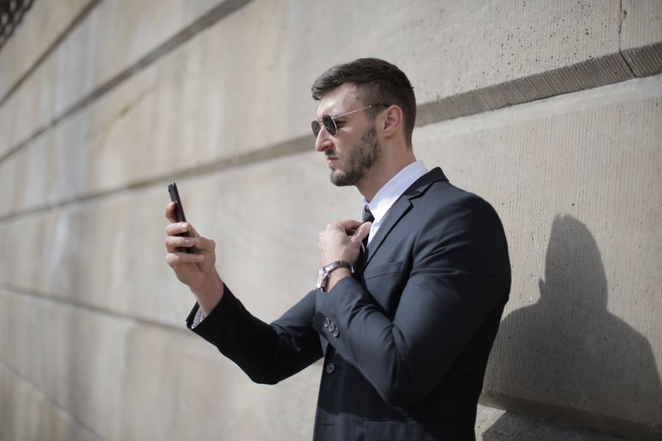A businessman adjusting tie while using smartphone outdoors, wearing sunglasses and a suit.
