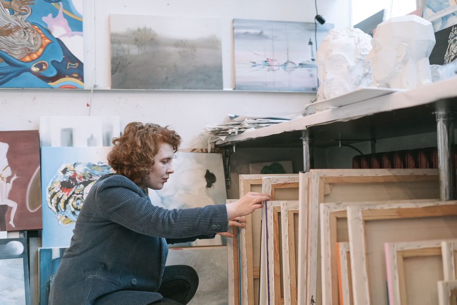 A focused artist organizes canvases in a cluttered art studio full of diverse artworks.
