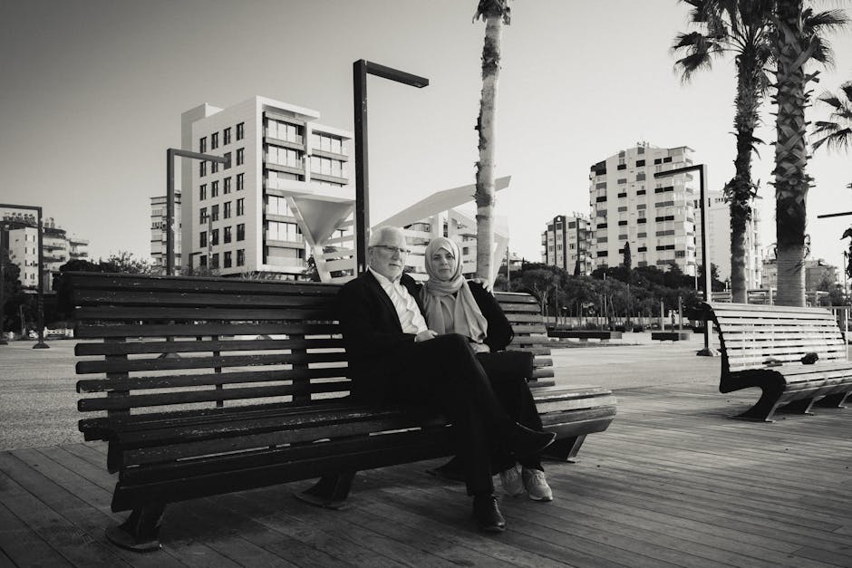 A grayscale image of a couple sitting together on a bench with urban architecture in the background.