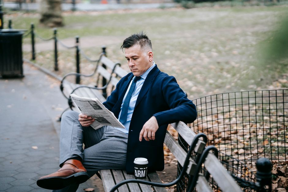Man in formal attire reading a newspaper on a park bench with a coffee cup.