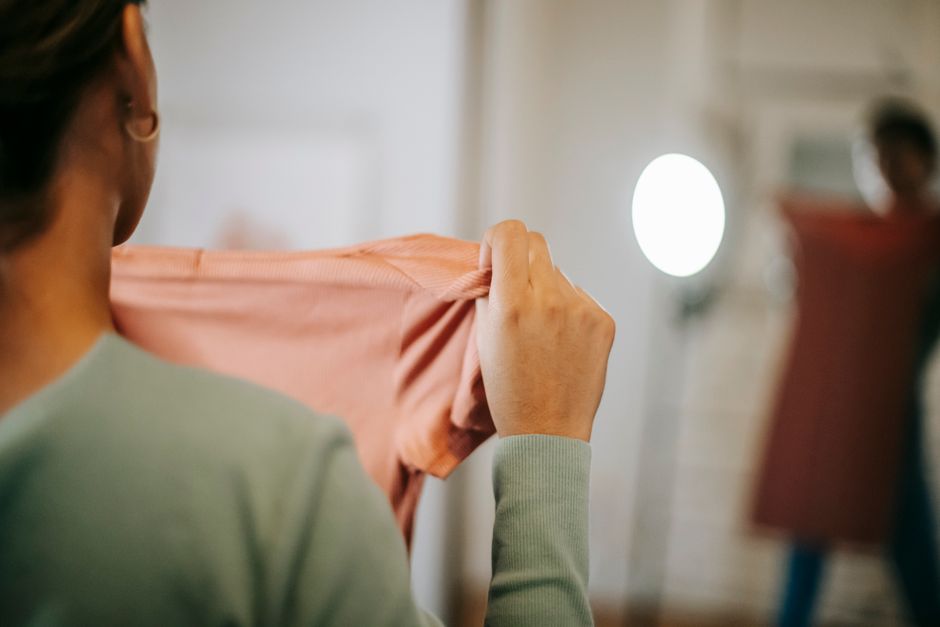 Back view of unrecognizable female in casual clothes showing pink dress while selecting clothes and looking in mirror while standing in light room at home