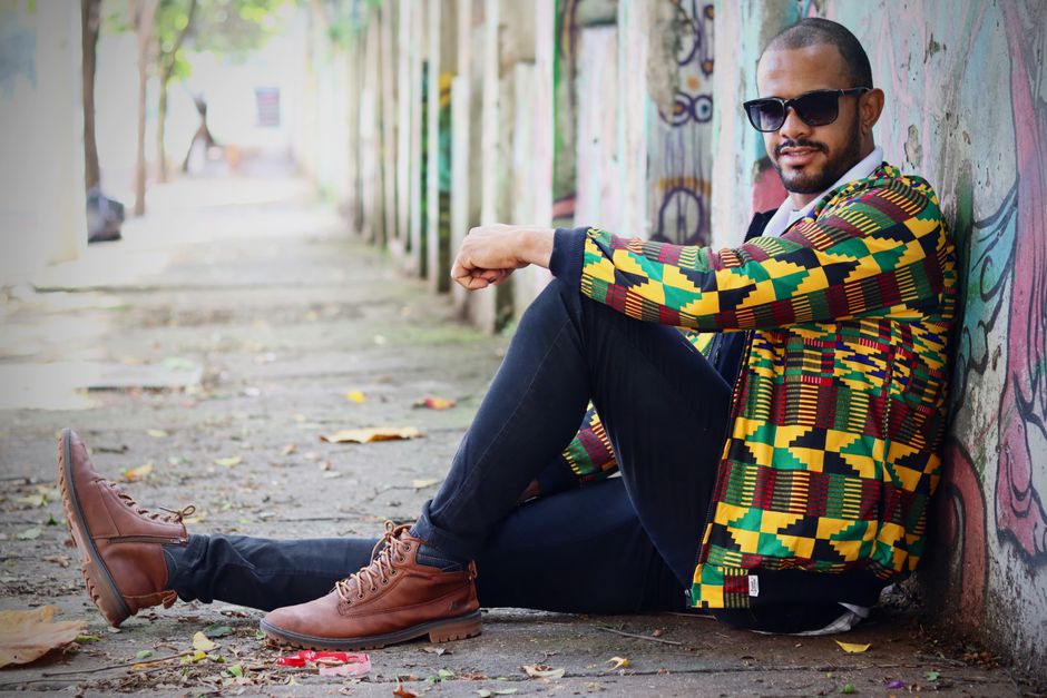 Man wearing African Kente jacket in urban outdoors, seated on sidewalk.