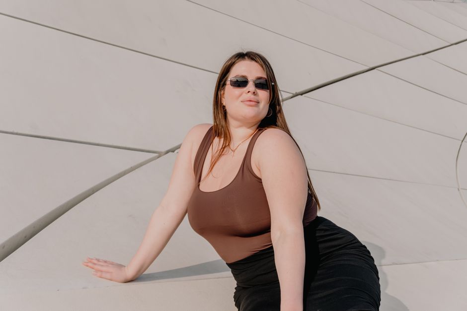 Confident woman wearing sunglasses poses against modern architectural backdrop in daylight.