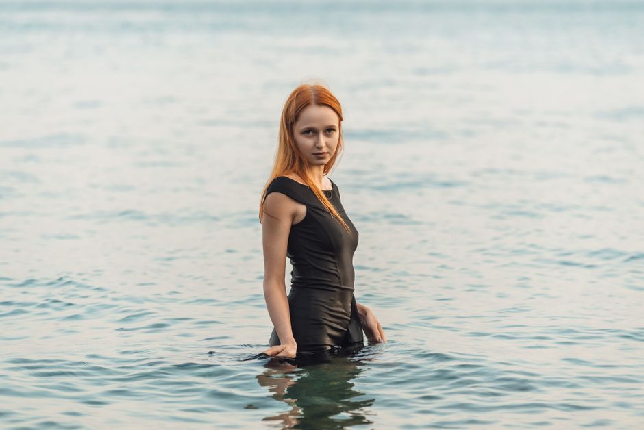 A young woman with long hair standing in the sea, wearing a black dress on a calm day.