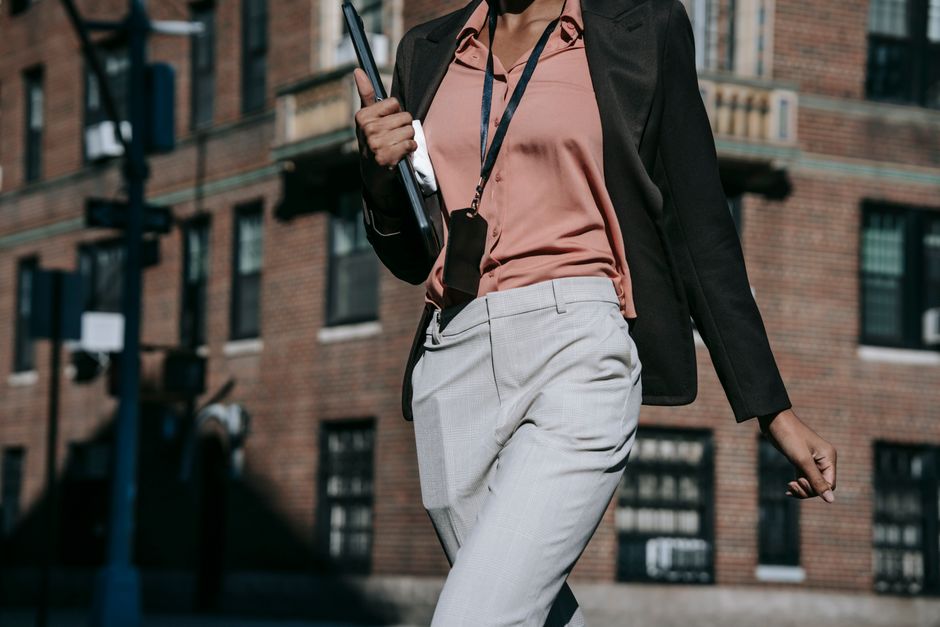 Crop anonymous young trendy black female entrepreneur in elegant clothes and name tag walking with laptop in hand on sunny day in city