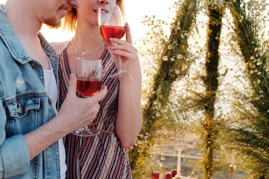 Romantic couple holding wine glasses outdoors amidst floral decorations at sunset.