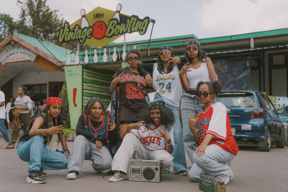 A group of stylish young adults pose in vintage sports jerseys outside a bowling alley.