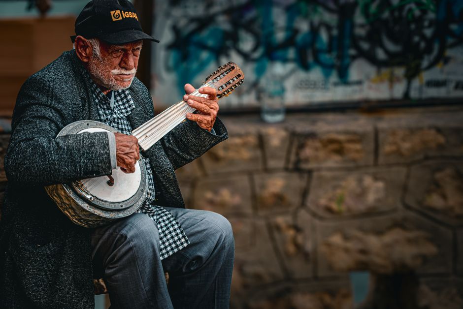 A senior man plays a mandolin outdoors, against a graffiti backdrop, showcasing street music culture.