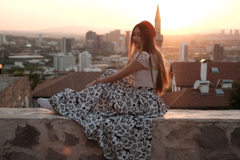 Asian woman in patterned dress sitting on a rooftop at sunset with an urban cityscape view.