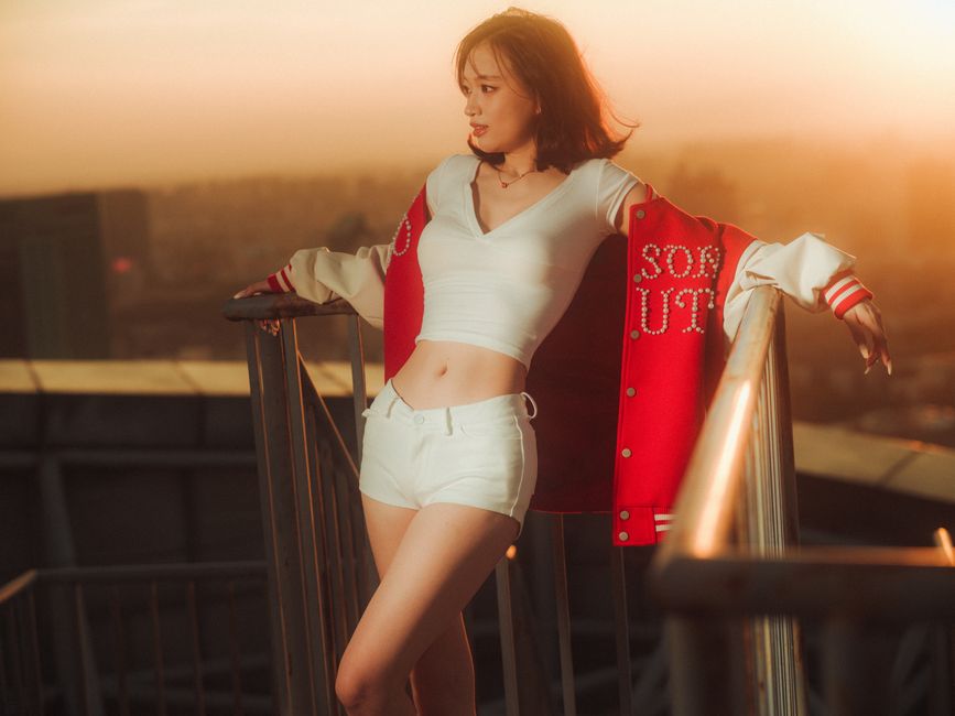 Framed by golden sunset hues, a stylish woman in a red jacket poses confidently on a rooftop.